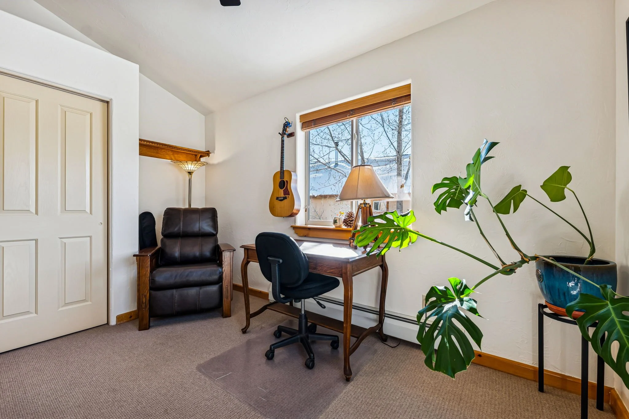 Home office with a wooden desk and black office chair by a window, a guitar hanging on the wall, a leather armchair, a floor lamp, a potted Monstera plant, and winter snow visible outside.