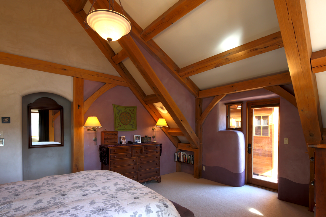 A cozy bedroom with wooden beams on the ceiling, a bed with a floral duvet cover, a dresser with framed photos and decorative items, a wall mirror, a small bookshelf, a purple accent wall, a window, and a balcony door.