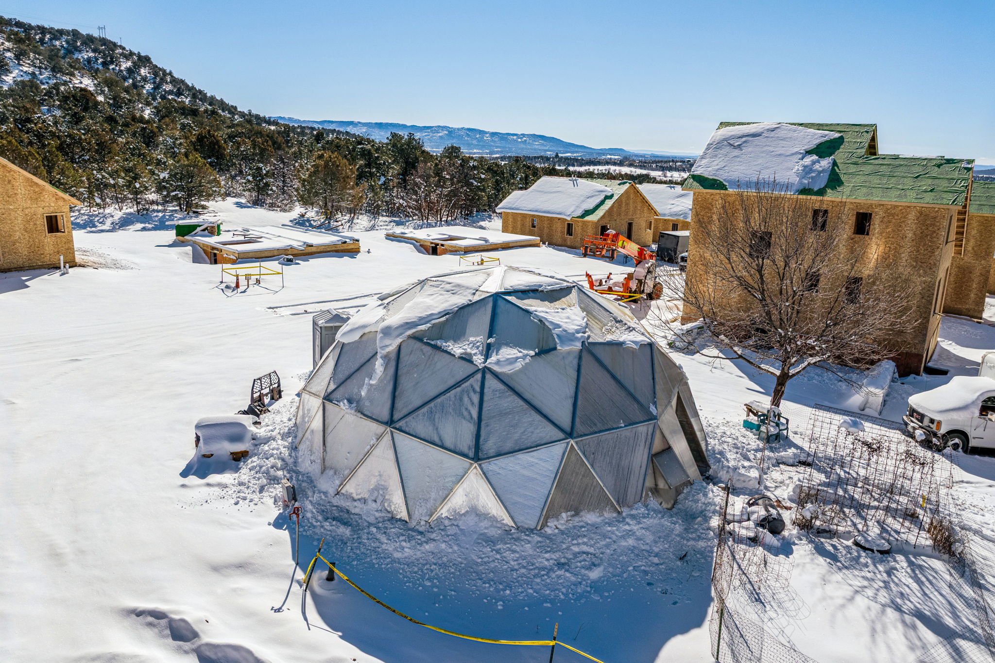A snowy backyard with a geodesic dome greenhouse, surrounded by unfinished houses with green roofs, trees, and equipment, under a clear blue sky.