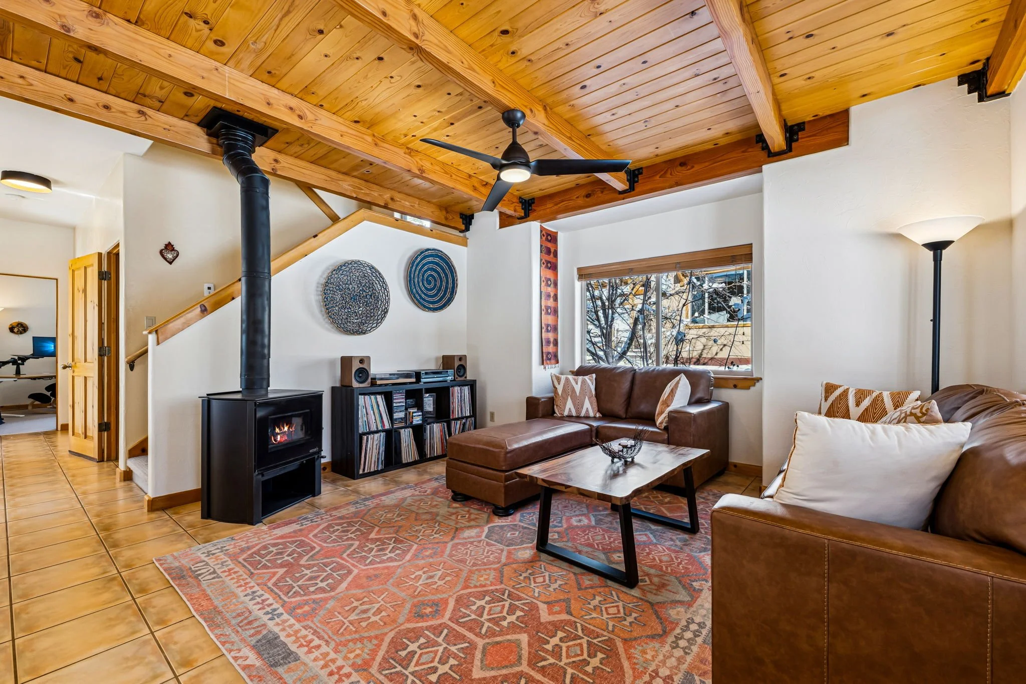 Living room with wooden ceiling, leather sofas, a coffee table, a fireplace, a window with a view of trees, and vinyl records and artwork on the wall.