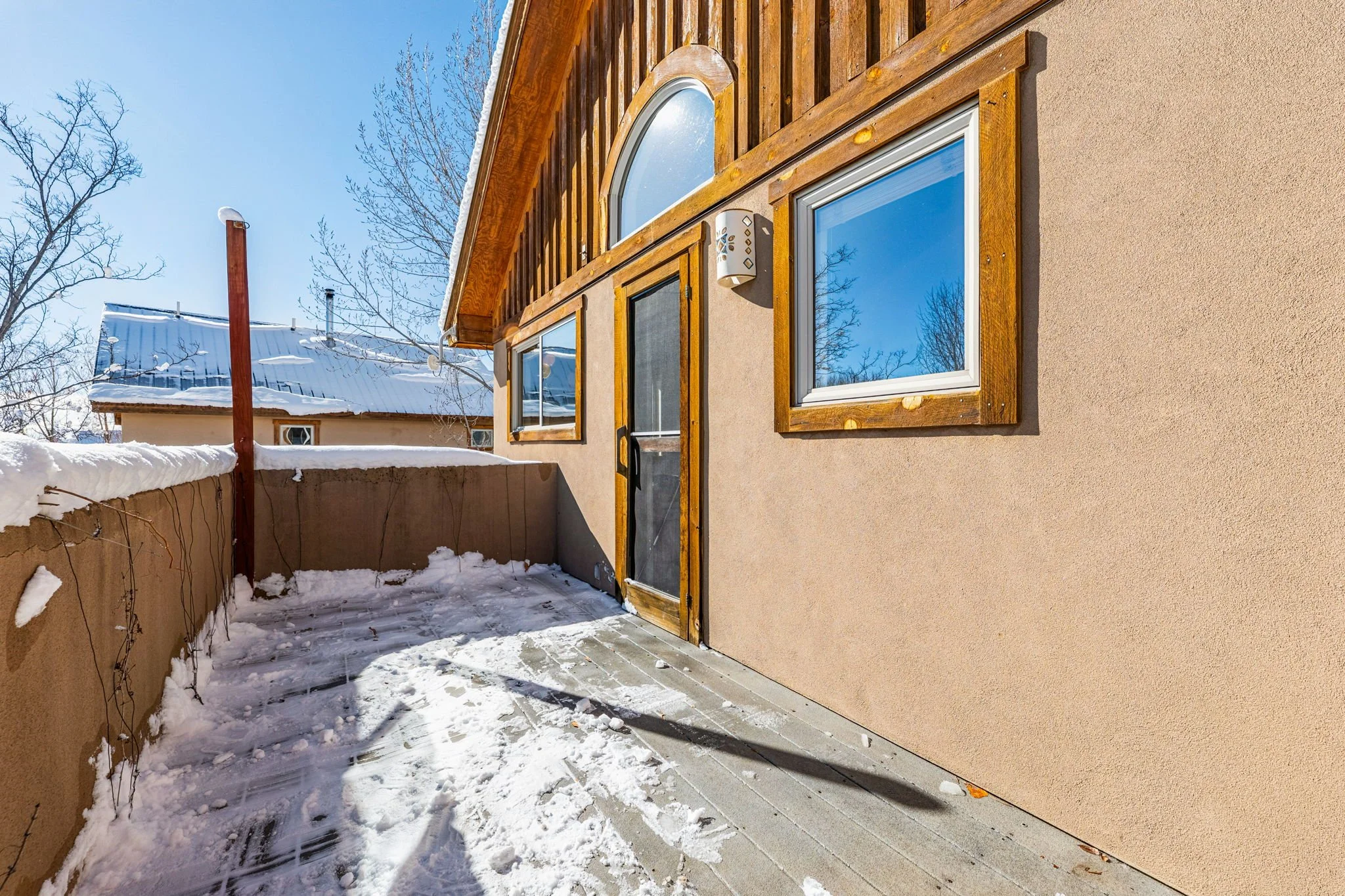 Image of a snow-covered balcony or terrace area attached to a beige house, with a wooden door and three windows, some with wood trim, and snow on the ground and railing. There are leafless trees and neighboring buildings in the background under a cle