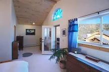 Interior view of a bedroom with a large window, blue curtains, a wooden dresser, a potted plant, and a doorway leading to another room.