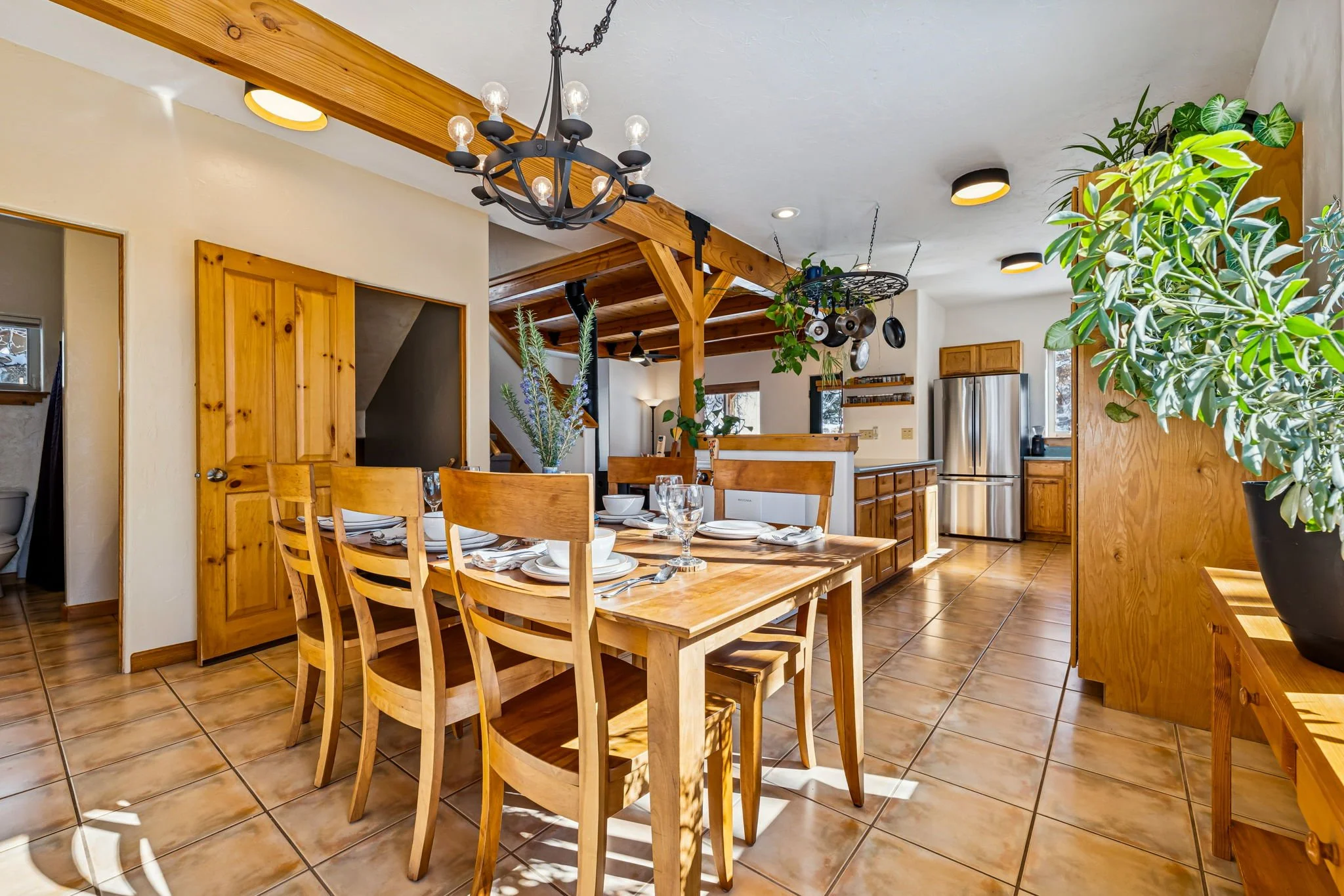 Dining area with a wooden dining table set for six, with white plates, glasses, and napkins, in a kitchen with wooden cabinets, stainless steel refrigerator, and fresh green plants, illuminated by ceiling lights and a chandelier.