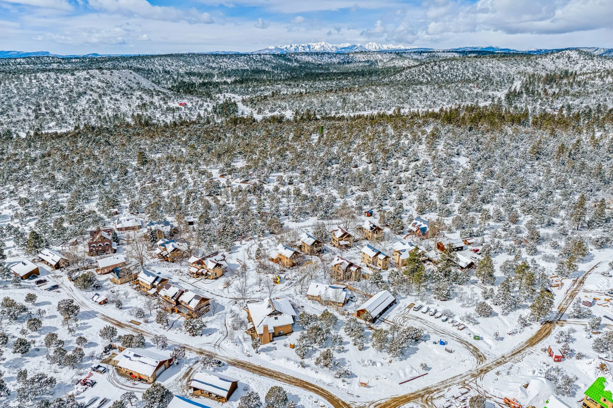 Aerial view of a snow-covered mountain village with houses, trees, and snow-covered roads in a forested mountainous region.