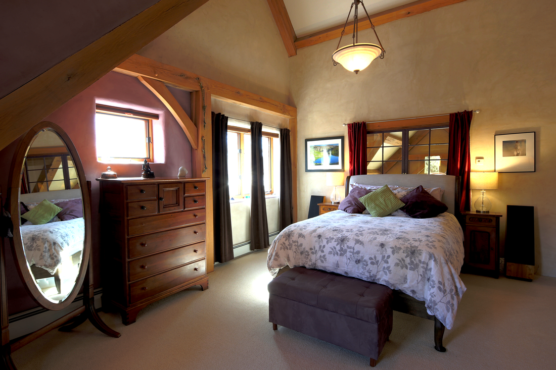 A cozy bedroom with a large bed, nightstands, and a dresser, illuminated by natural and artificial light, featuring arched windows, curtains, and framed artwork.