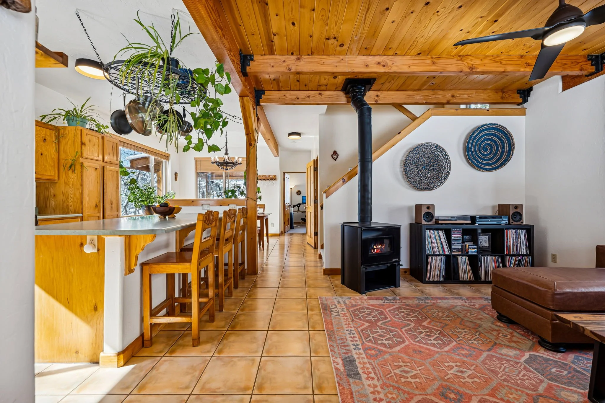 Living room with wood-paneled ceiling, a wood stove with pipe, a bookshelf with vinyl records, a brown leather ottoman, wall art, and a tiled floor.