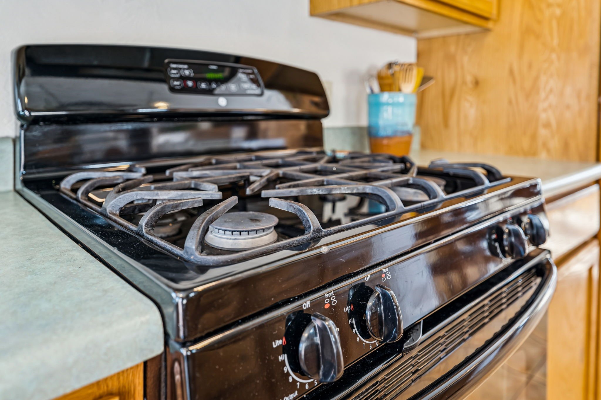 Close-up of a black gas stove with four burners in a kitchen. The stove has control knobs below, with one knob turned to a low setting. There is a light-colored countertop and wooden cabinets in the background.
