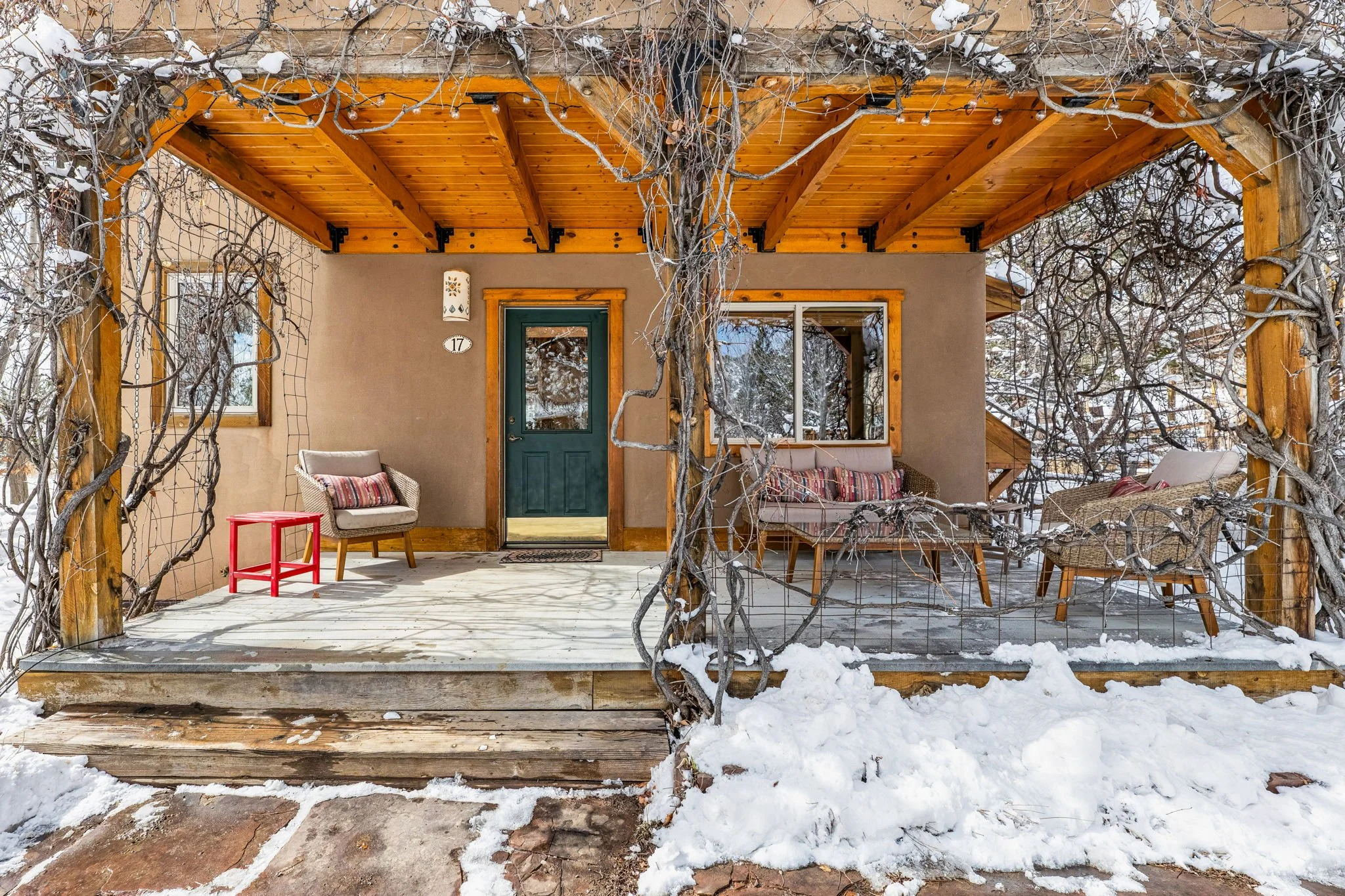 Front porch of a house with snow on the ground, featuring wooden beams, a small seating area with wicker chairs, a red side table, and trees with bare branches.