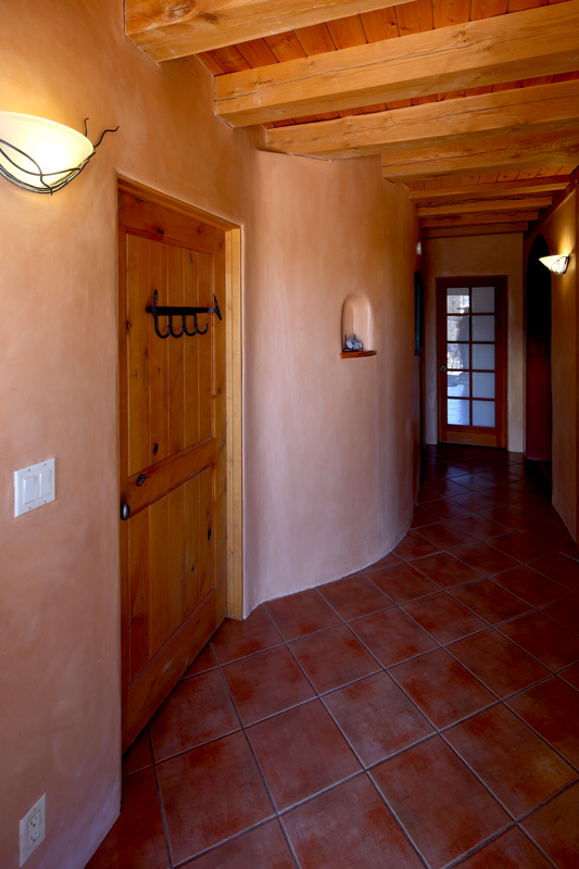 Interior hallway with brick red tiled floor, curved wall with a small shelf, wooden door with hooks, and wooden ceiling beams, with wall-mounted lights.