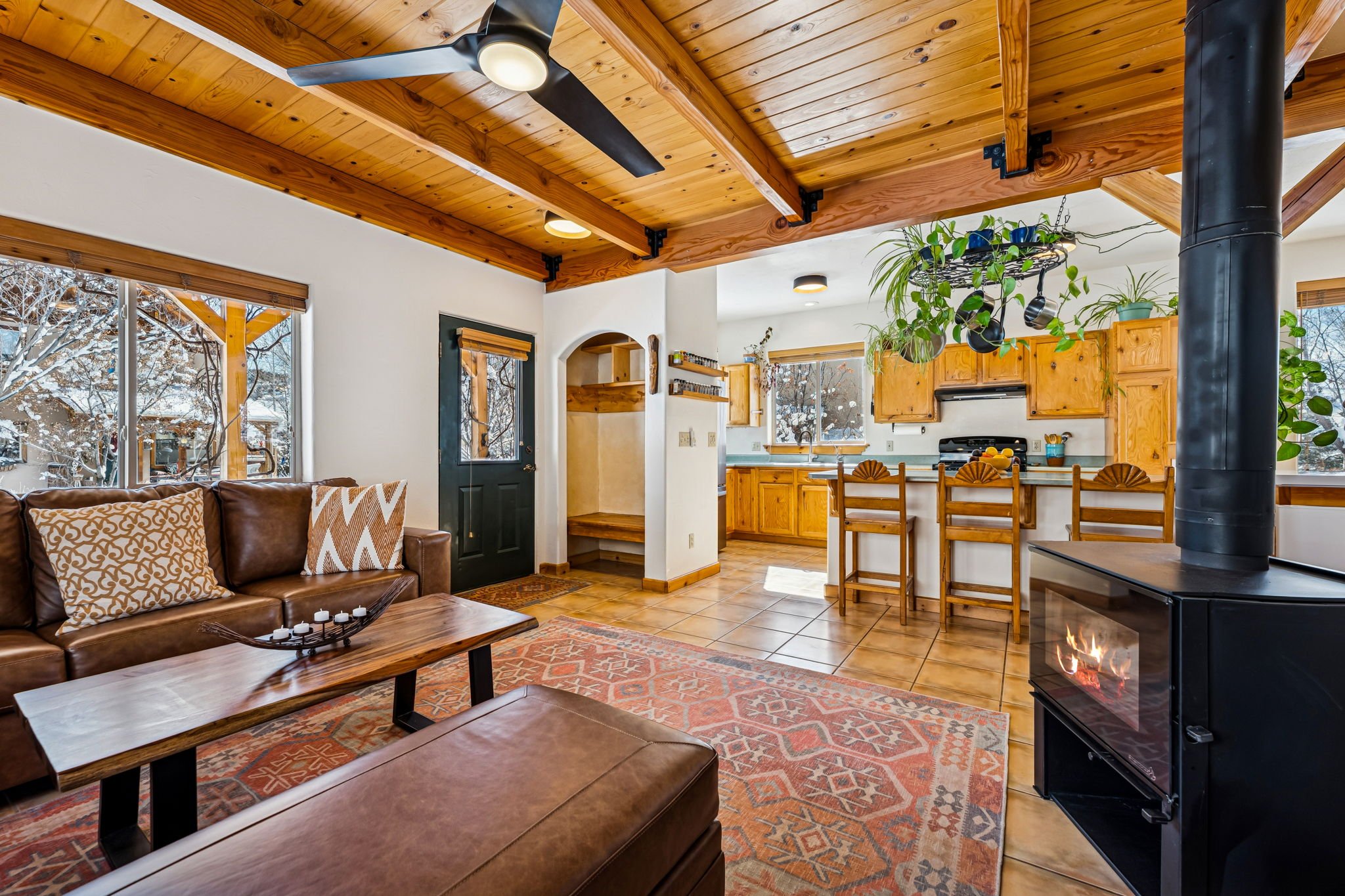 Living room and kitchen with wooden accents, a leather couch with decorative pillows, a wooden coffee table, a black wood stove with a visible flame, a ceiling fan, and large windows showing snow outside