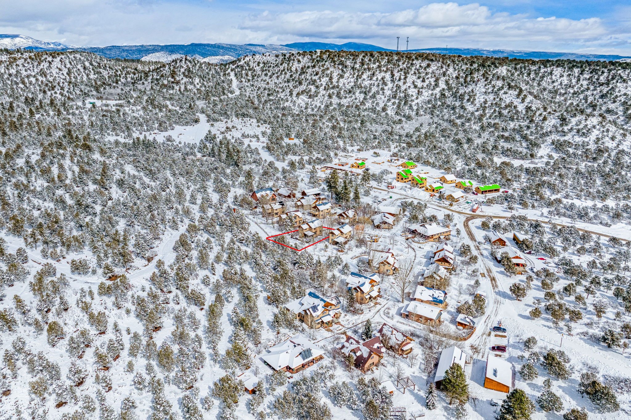 A snowy village with houses and trees covered in snow, surrounded by forested hills and mountains under a cloudy sky.