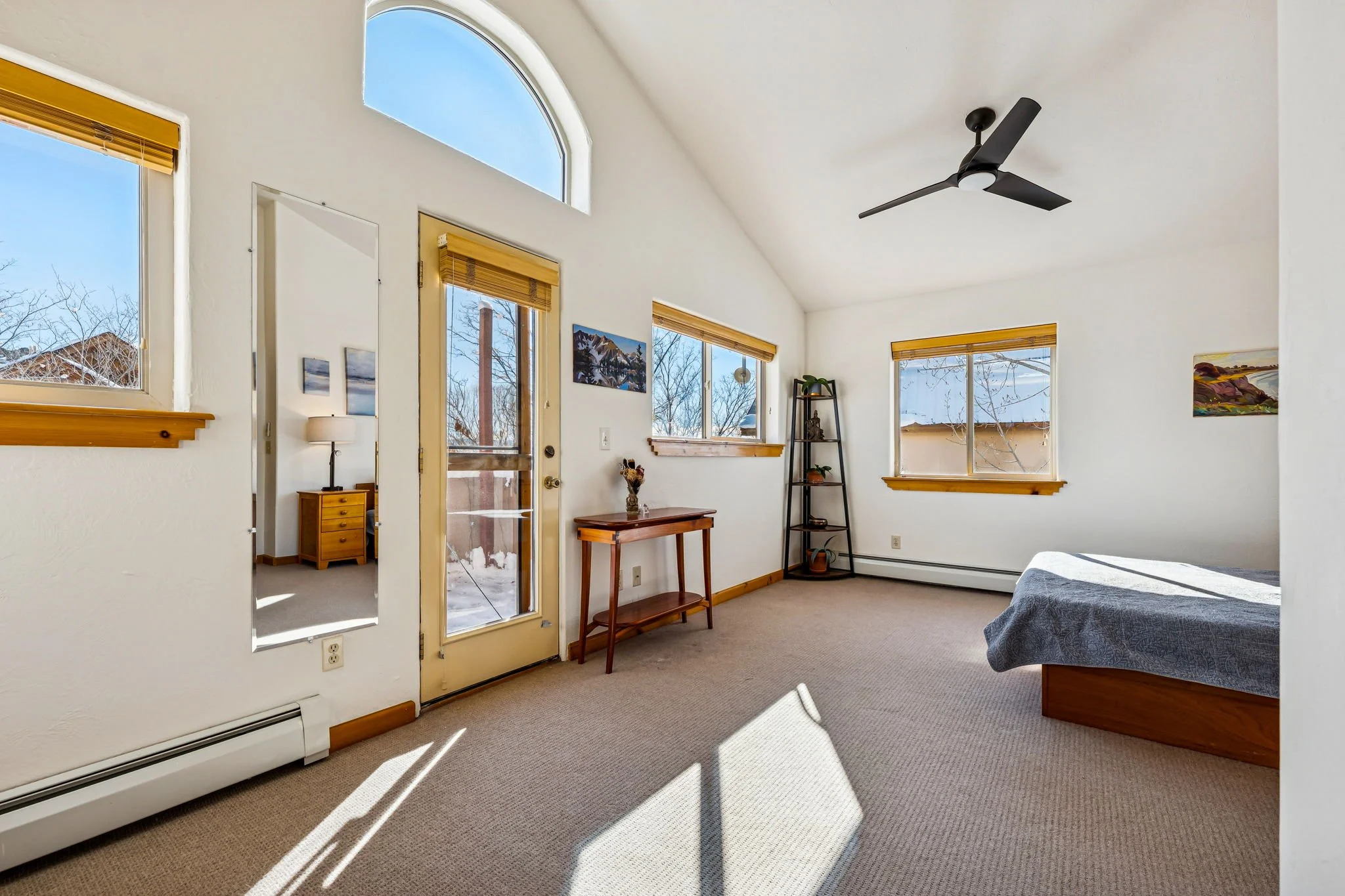 Bright bedroom with white walls, multiple windows with yellow blinds, and a view of snow outside. Contains a wooden bed with a blue blanket, a black metal shelf with decor, a small wooden table, and a ceiling fan.