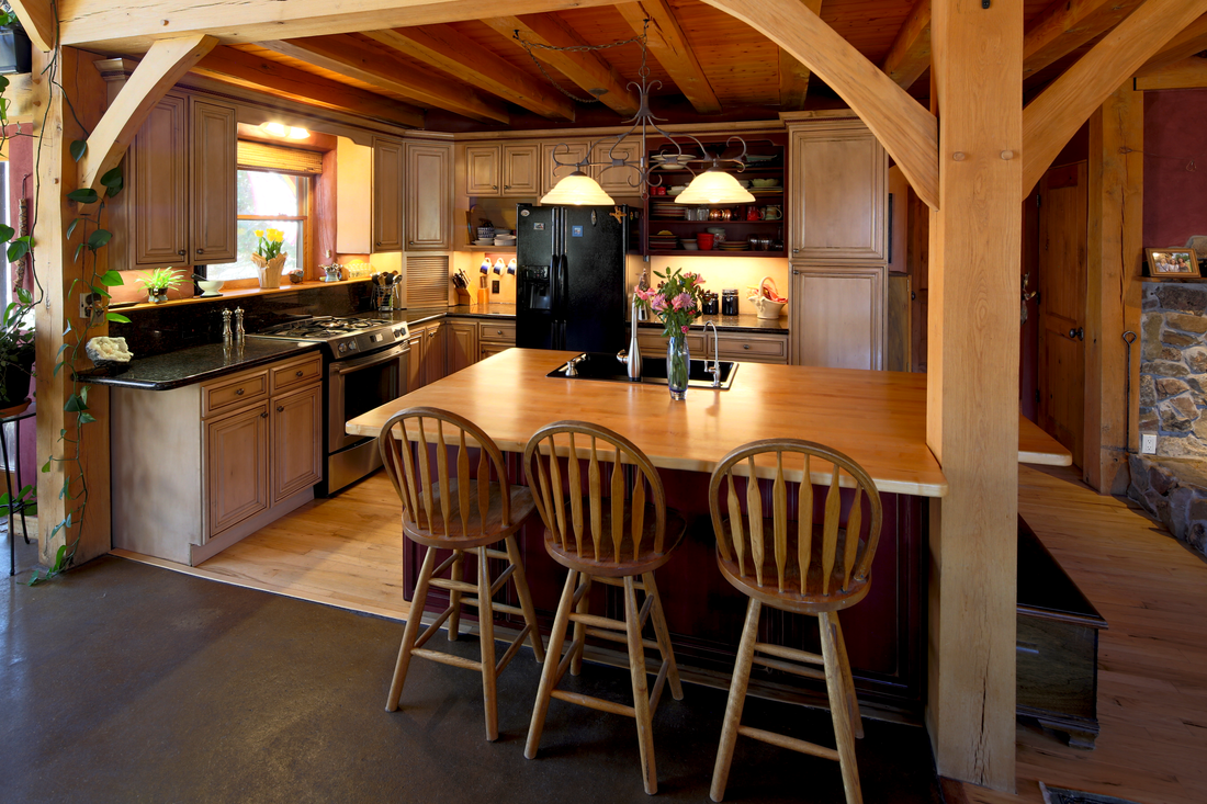 Kitchen with wooden cabinets, black refrigerator, black stove, wooden island with three chairs, window with plants, and wooden ceiling beams.