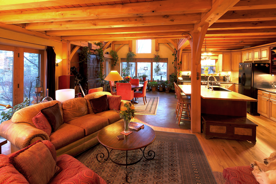 Open-concept living room and kitchen with wooden beams, a sofa, armchairs, a coffee table, a dining table with chairs, and a kitchen with wooden cabinets and black refrigerator, illuminated by natural light from large windows.
