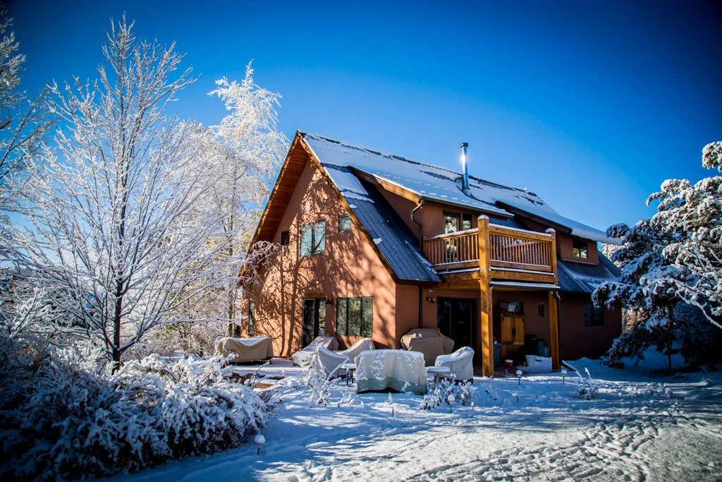 A two-story house surrounded by snow-covered trees and bushes, with outdoor furniture covered in snow, under a clear blue sky