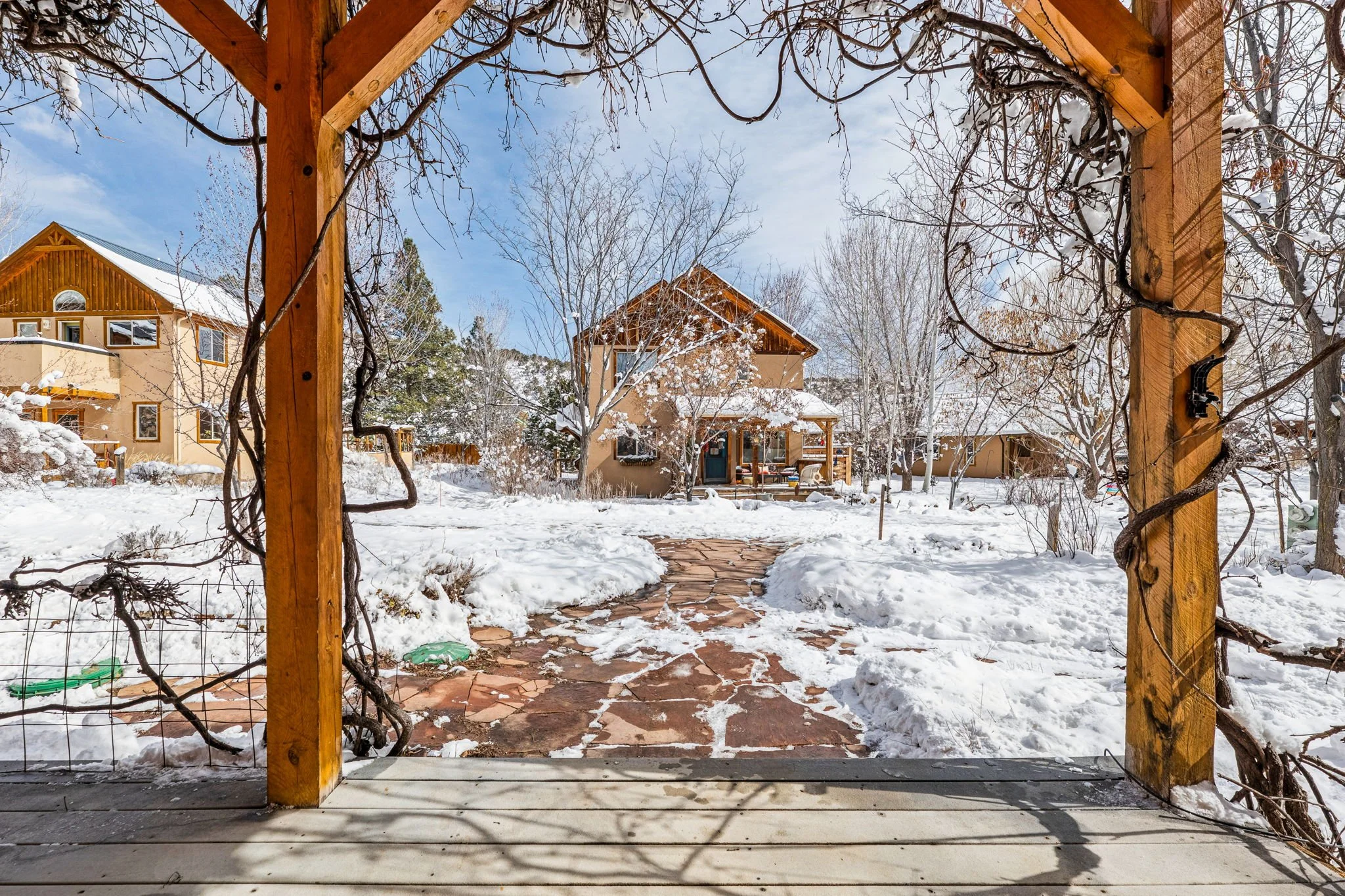 View from a wooden porch looking out onto a snowy backyard and neighboring houses with snow-covered roofs, leafless trees, and a blue sky.