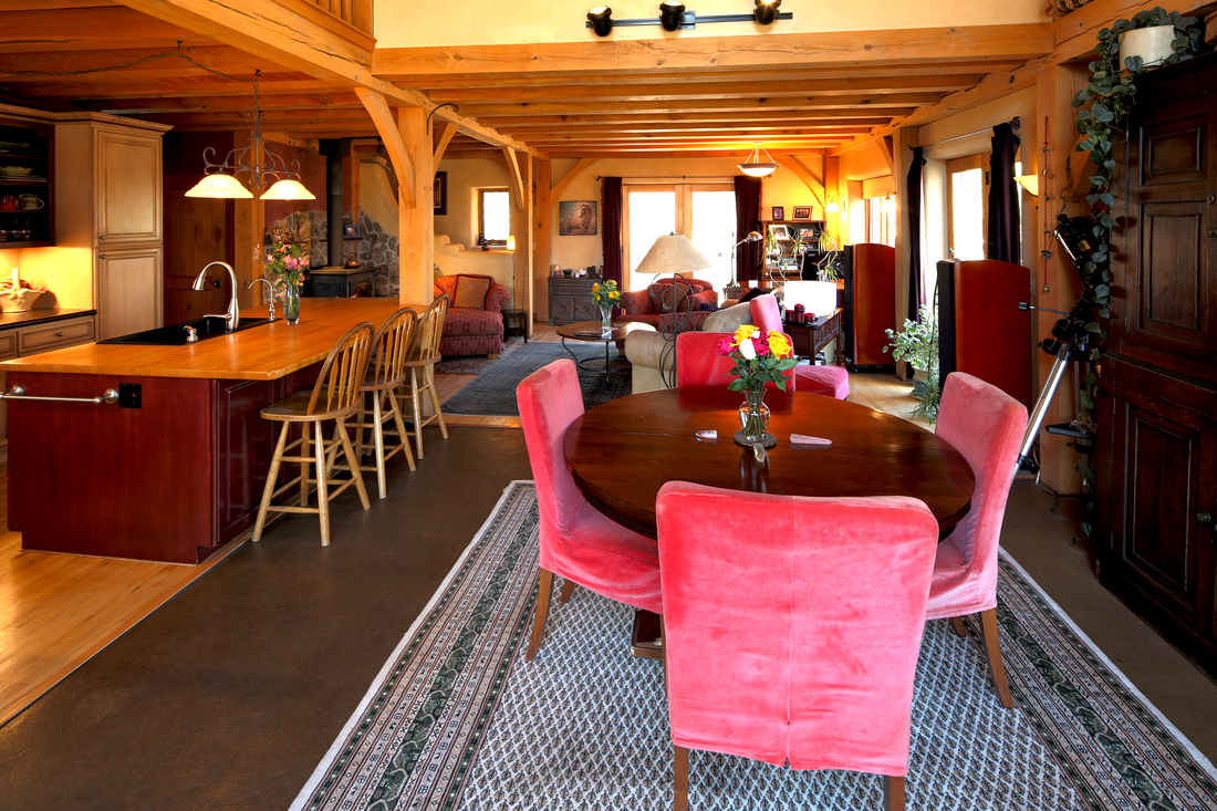 Living room with wooden beams, a round dining table with pink velvet chairs, a kitchen area with a breakfast bar and wooden stools, and a cozy seating area with armchairs, lamps, and large windows.