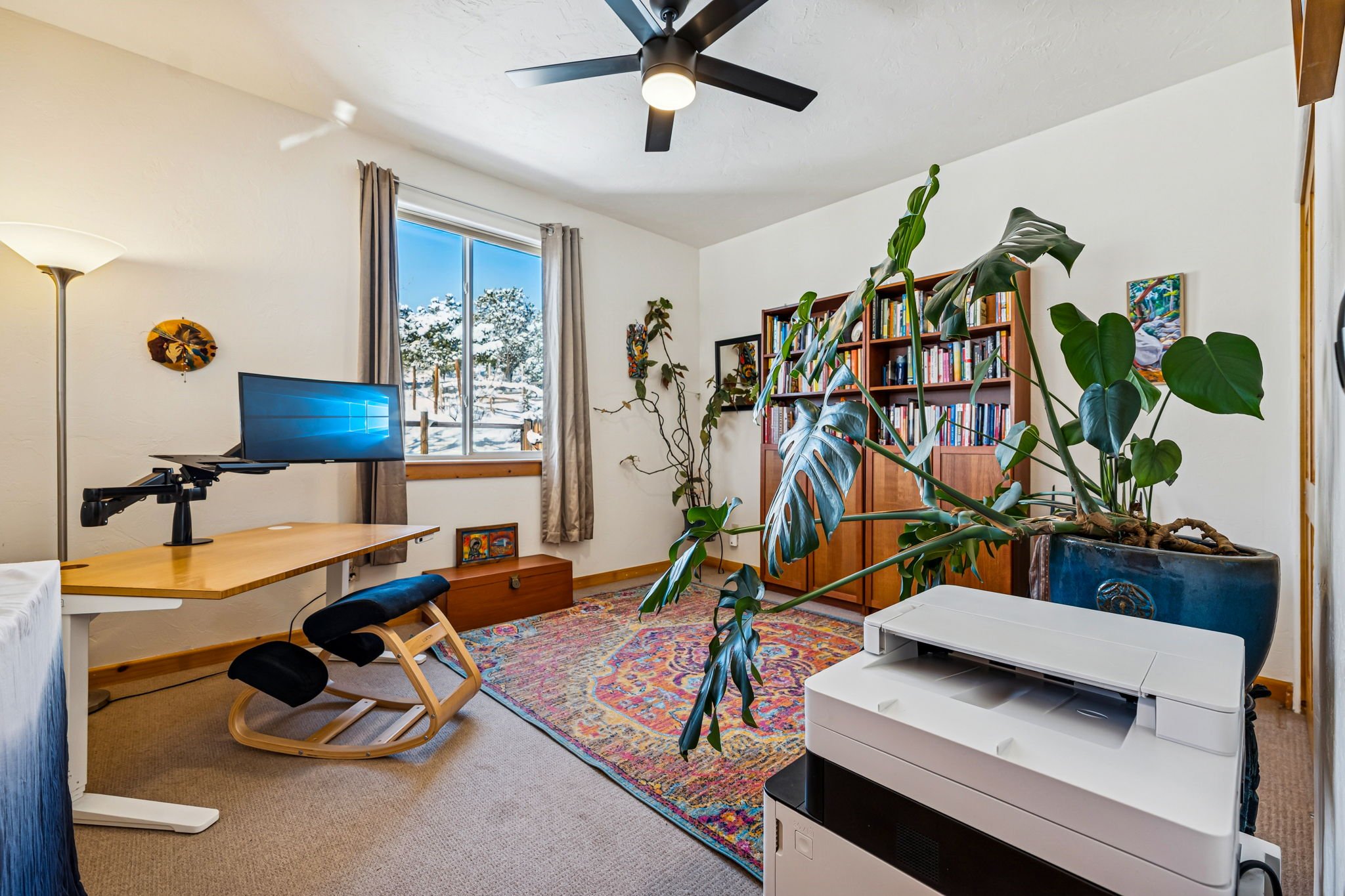 A home office or study room with a window showing snow outside, a wooden desk with a monitor, a mauve-colored chair, a bookshelf filled with books, a large potted monstera plant, a colorful area rug, a standing lamp, and a white printer.