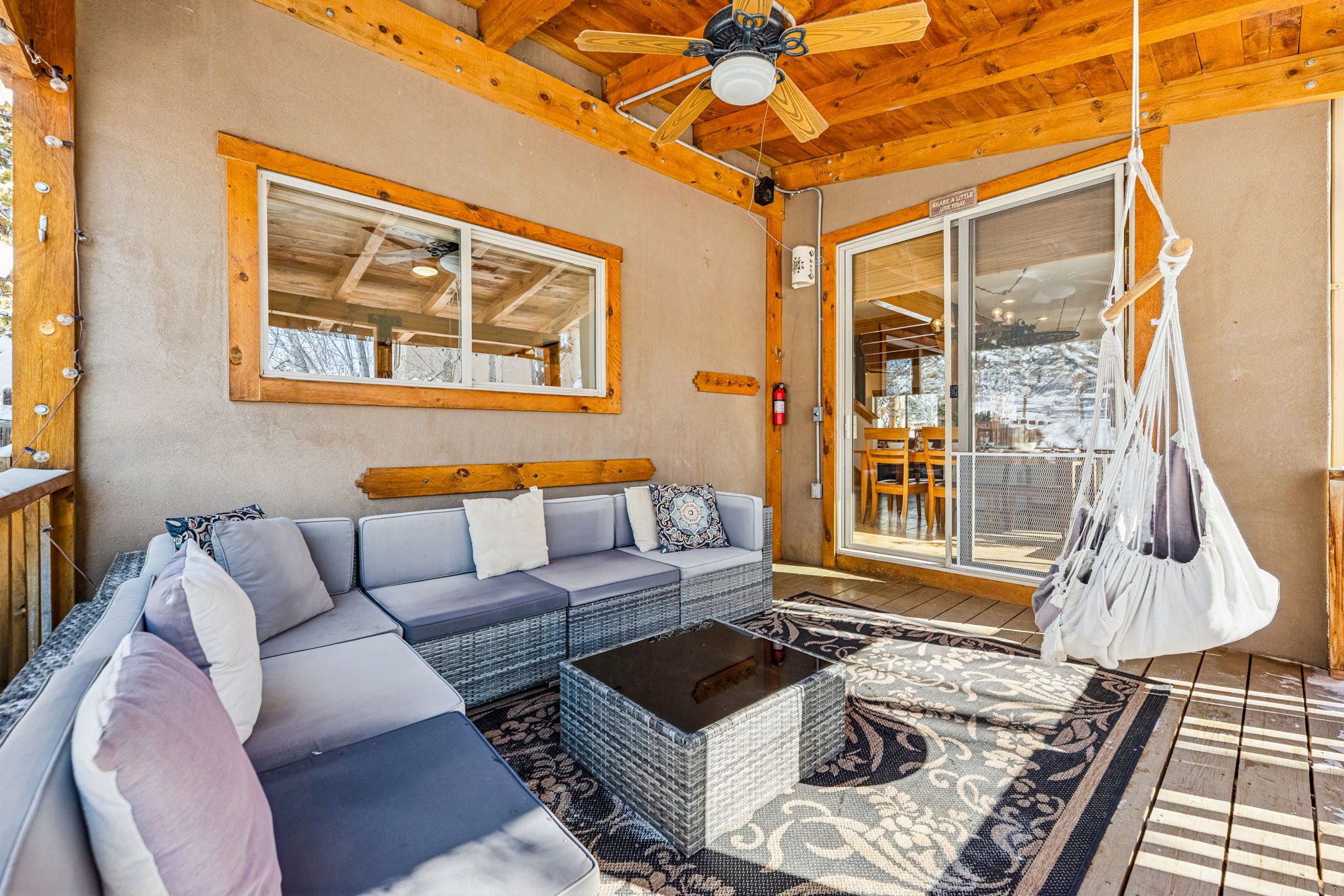 Sunlit outdoor patio with beige stucco walls, wooden ceiling with a ceiling fan, and sliding glass door. Features a gray wicker sectional sofa with pillows, an outdoor patterned rug, a black coffee table, and a hanging hammock chair.