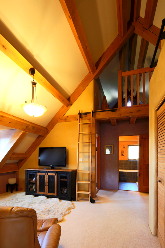 Interior of a living space with a high vaulted ceiling, exposed wooden beams, a small ladder, and a loft area. There is a television on a black wooden stand, a cream-colored shag area rug, and a leather chair in the foreground. A ceiling light illumi