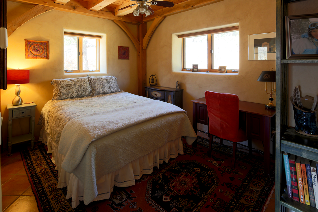 Cozy bedroom with a bed, wooden ceiling beams, two windows, a desk with a red chair, and decorative items.