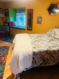 A bedroom with yellow walls, a bed with floral bedding, a window with green curtains, and a colorful rug on the wooden floor.