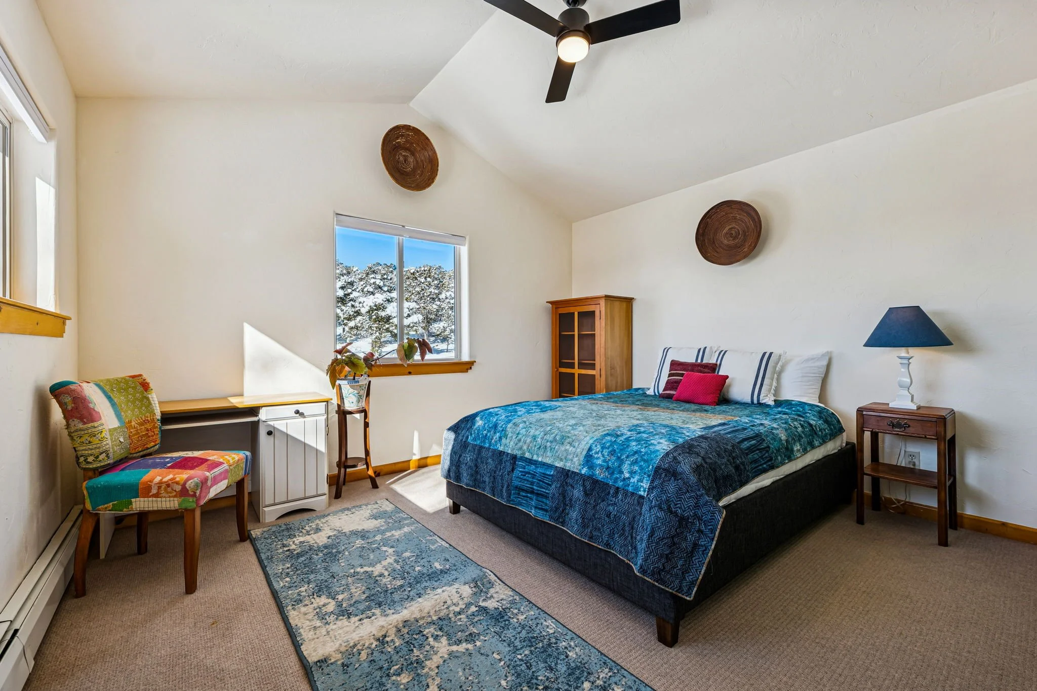 A cozy bedroom with a bed covered in a blue patterned quilt, a wooden nightstand with a blue lamp, a colorful patchwork chair, a window showing snowy trees, and a ceiling fan.