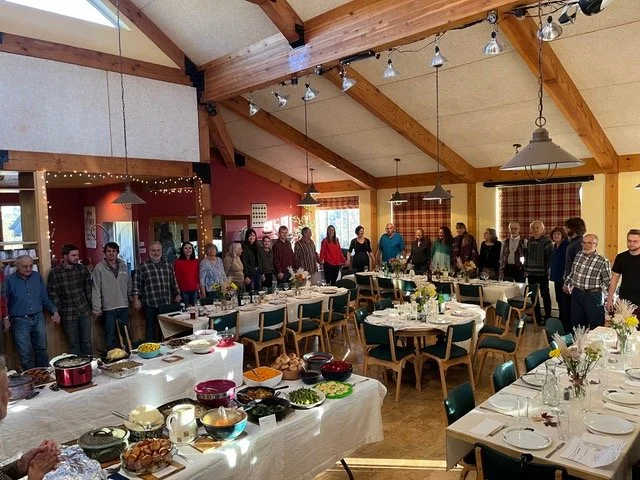 People standing in a large, decorated banquet hall with long tables set for a meal, serving dishes, and floral centerpieces.