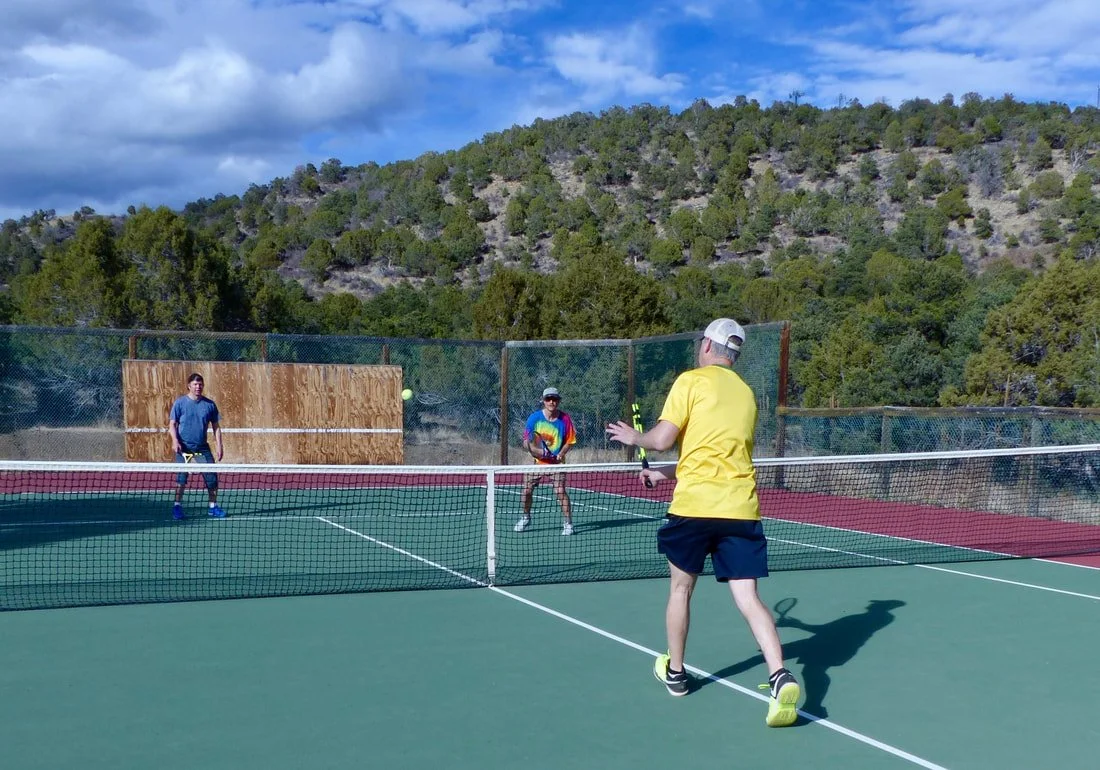 Three people playing pickleball on an outdoor court with mountains and trees in the background.