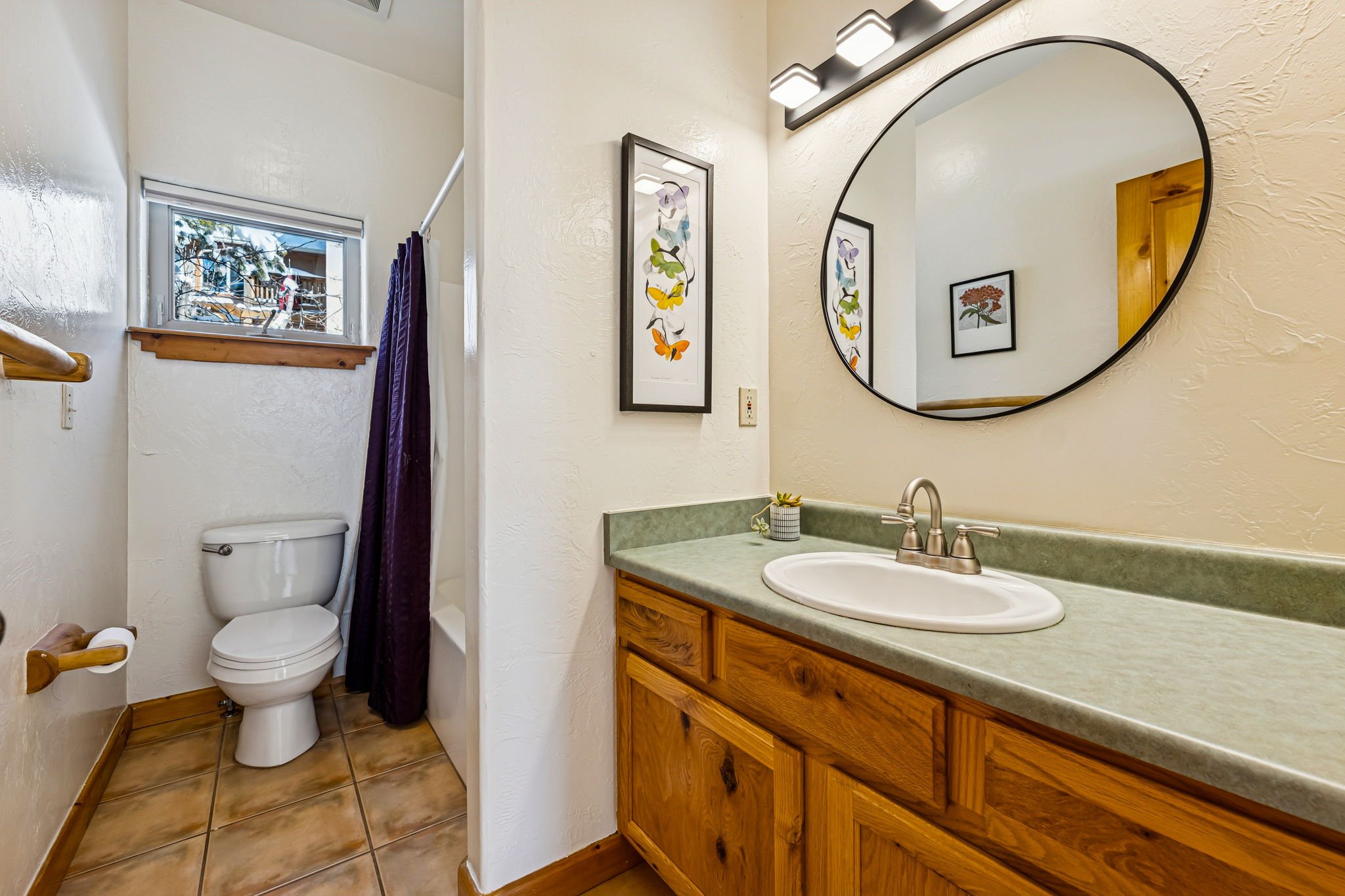 Bathroom with wooden vanity, green countertop, oval mirror, framed butterfly art, small window, toilet, shower stall with dark curtain.