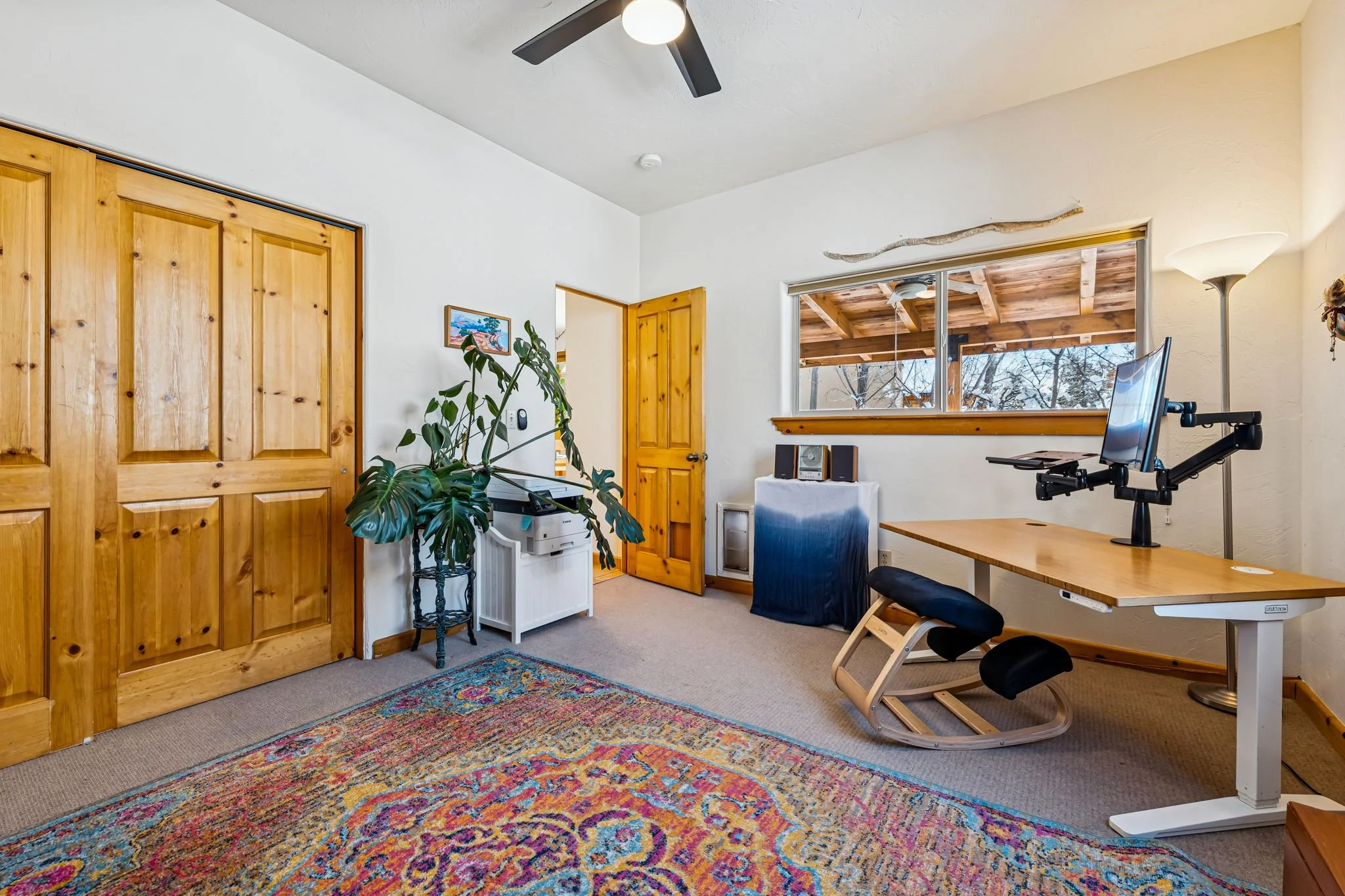 A cozy home office with a large wooden desk, ergonomic kneeling chair, window view of a wooden deck and trees, a standing lamp, a potted plant, and a colorful area rug on beige carpet.
