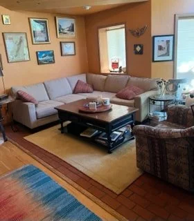 Living room with a beige sofa, pink pillows, and a dark coffee table, with framed pictures on orange-colored wall, a window, and a patterned armchair.
