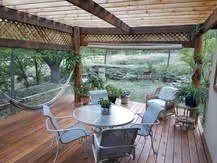 Covered wooden porch with a round table, six chairs, plants, and a view of a backyard with trees and grass.