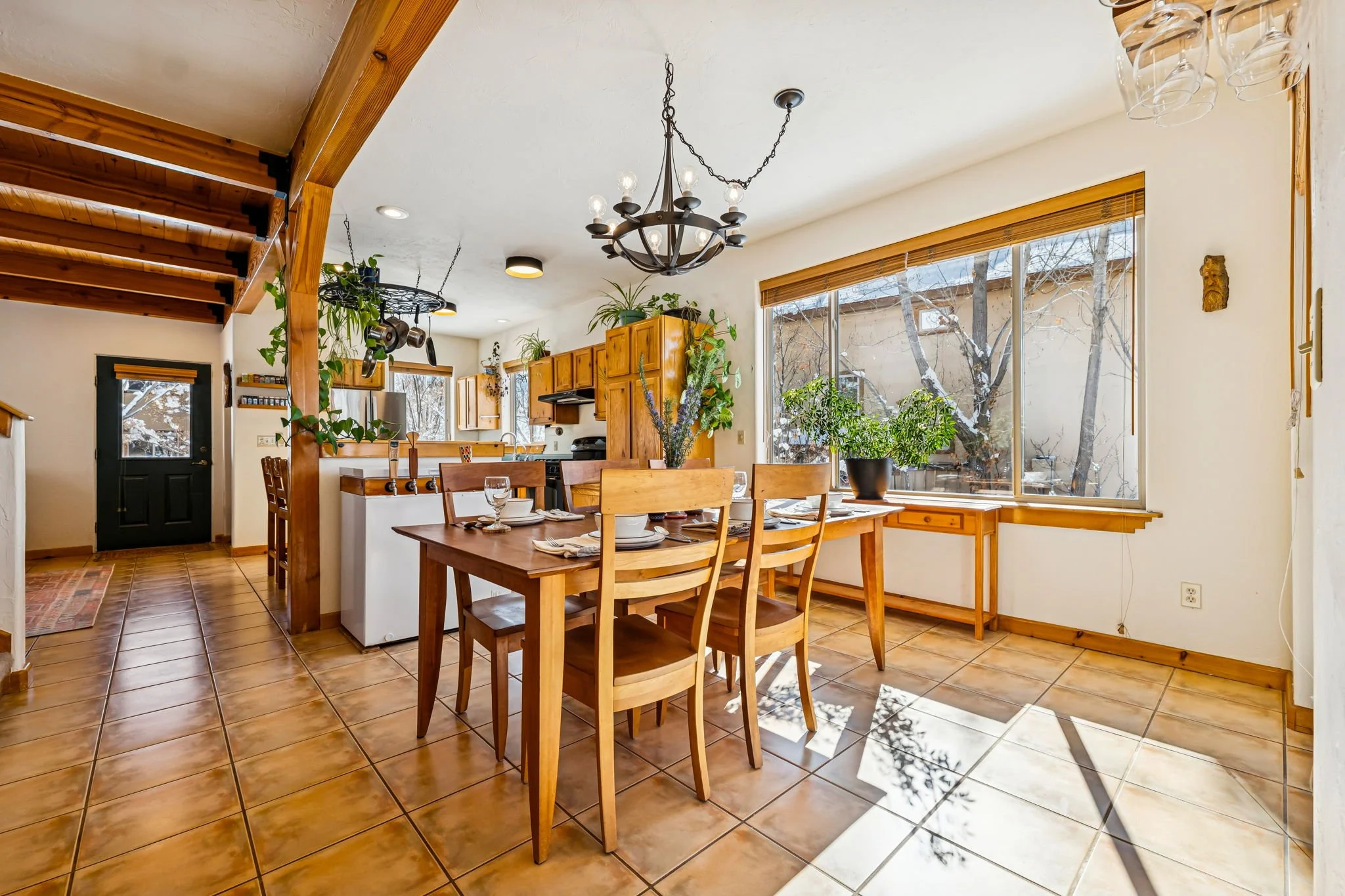 Dining area with a wooden table set for four, decorated with a vase, on a tile floor. Large window showing trees outside, and a kitchen with wooden cabinets in the background.