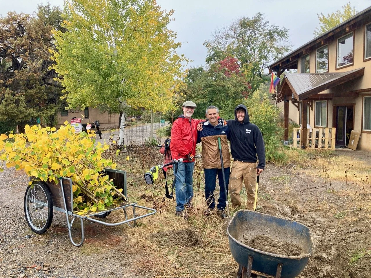 Three men standing outdoors with gardening tools, next to a wheelbarrow filled with dirt and a tree with yellow leaves, in front of a house with a flag and trees in the background.