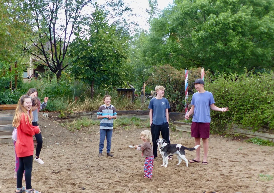 Children and a dog playing together outdoors in a yard with trees and bushes.