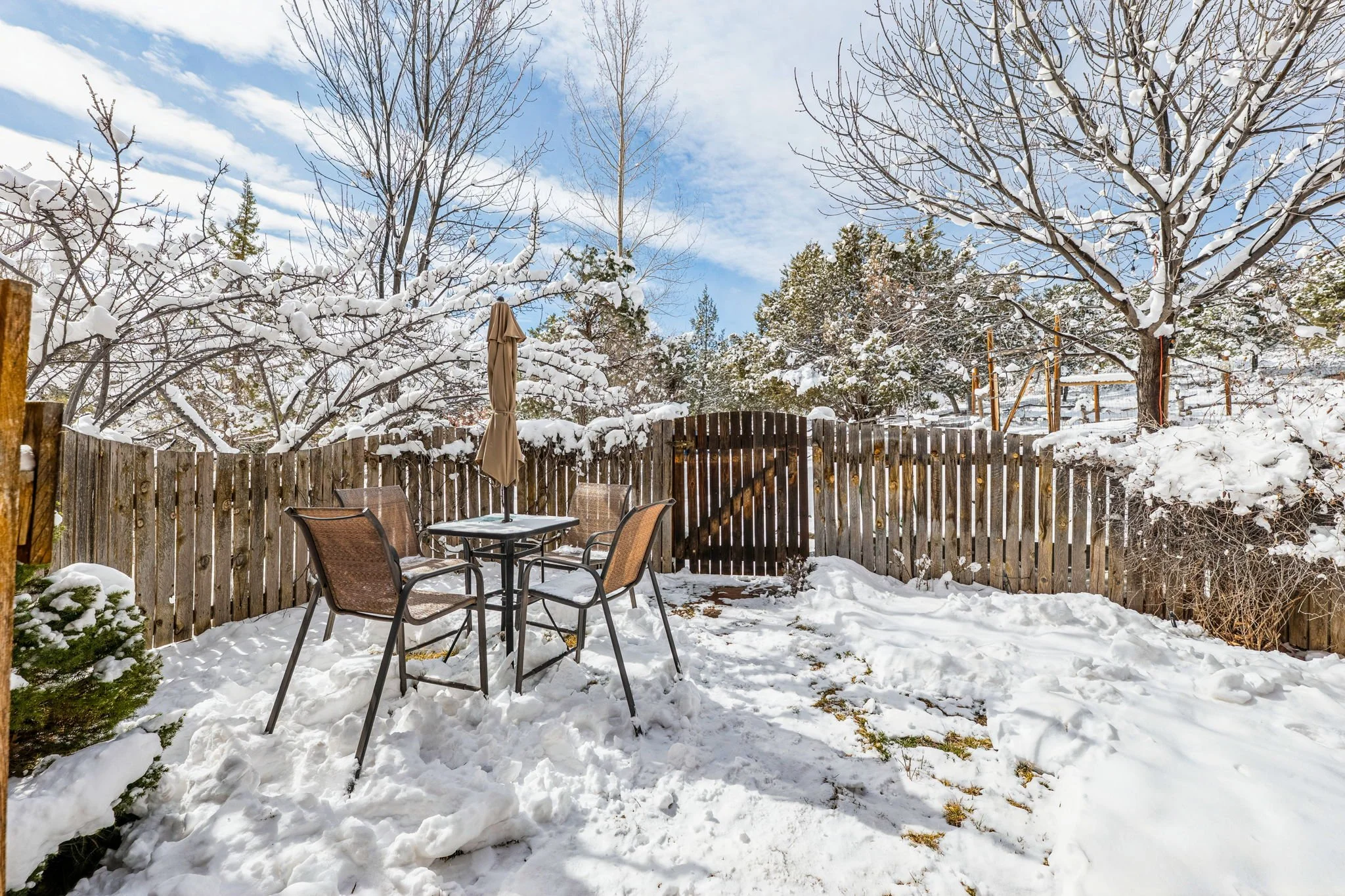 Snow-covered backyard patio with a glass table, four chairs, a closed umbrella, surrounded by a wooden fence and snow-laden trees under a partly cloudy sky.