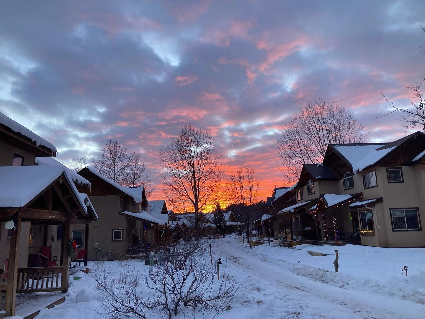 Snow-covered neighborhood street at sunset with colorful sky and bare trees.
