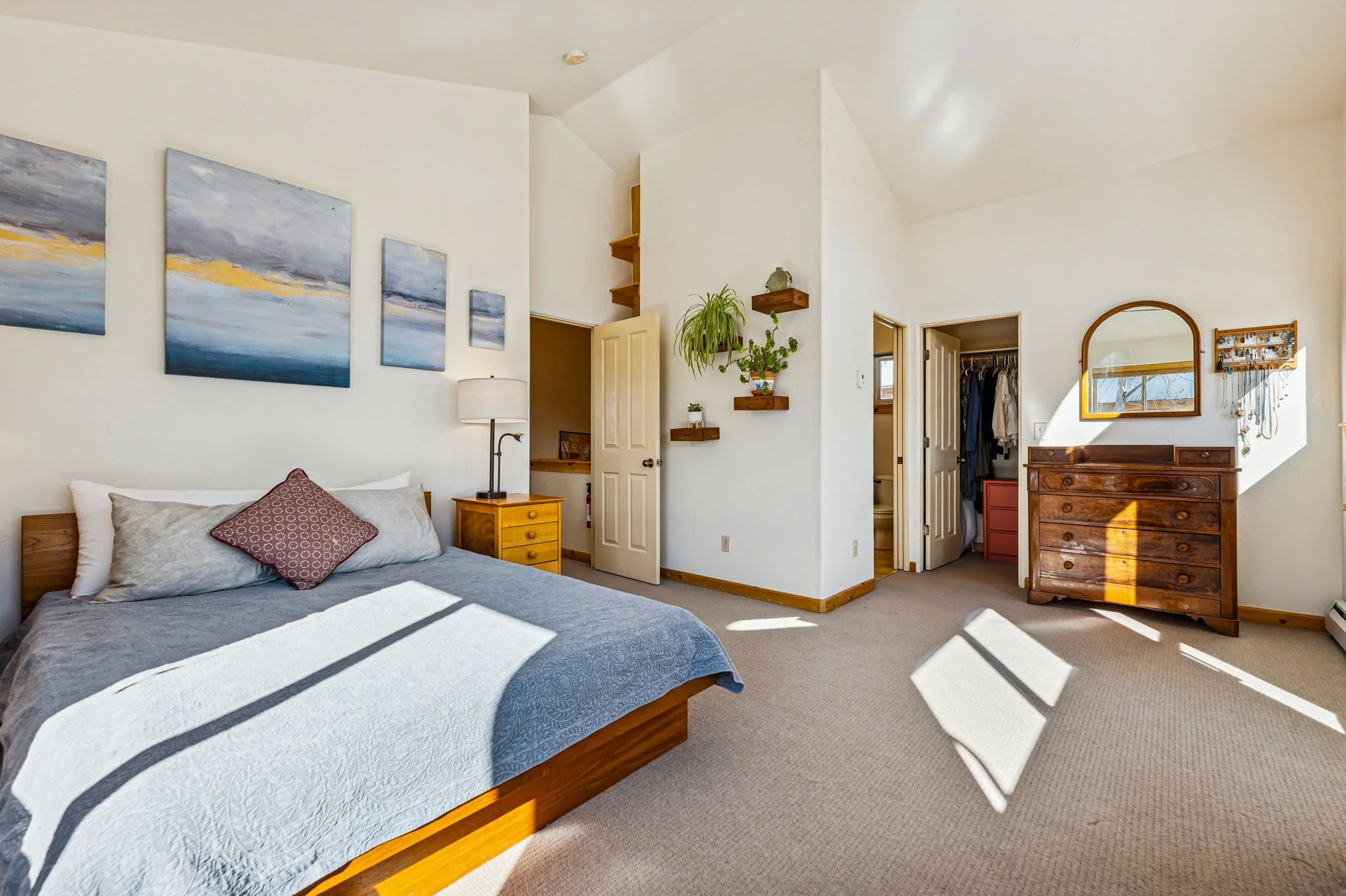 A bright bedroom with a bed, nightstand with lamp, and wall art. There are hanging plant shelves, a dresser with mirror, and two open doors leading to a walk-in closet and bathroom. Sunlight streams through windows, creating shadows on the carpeted f