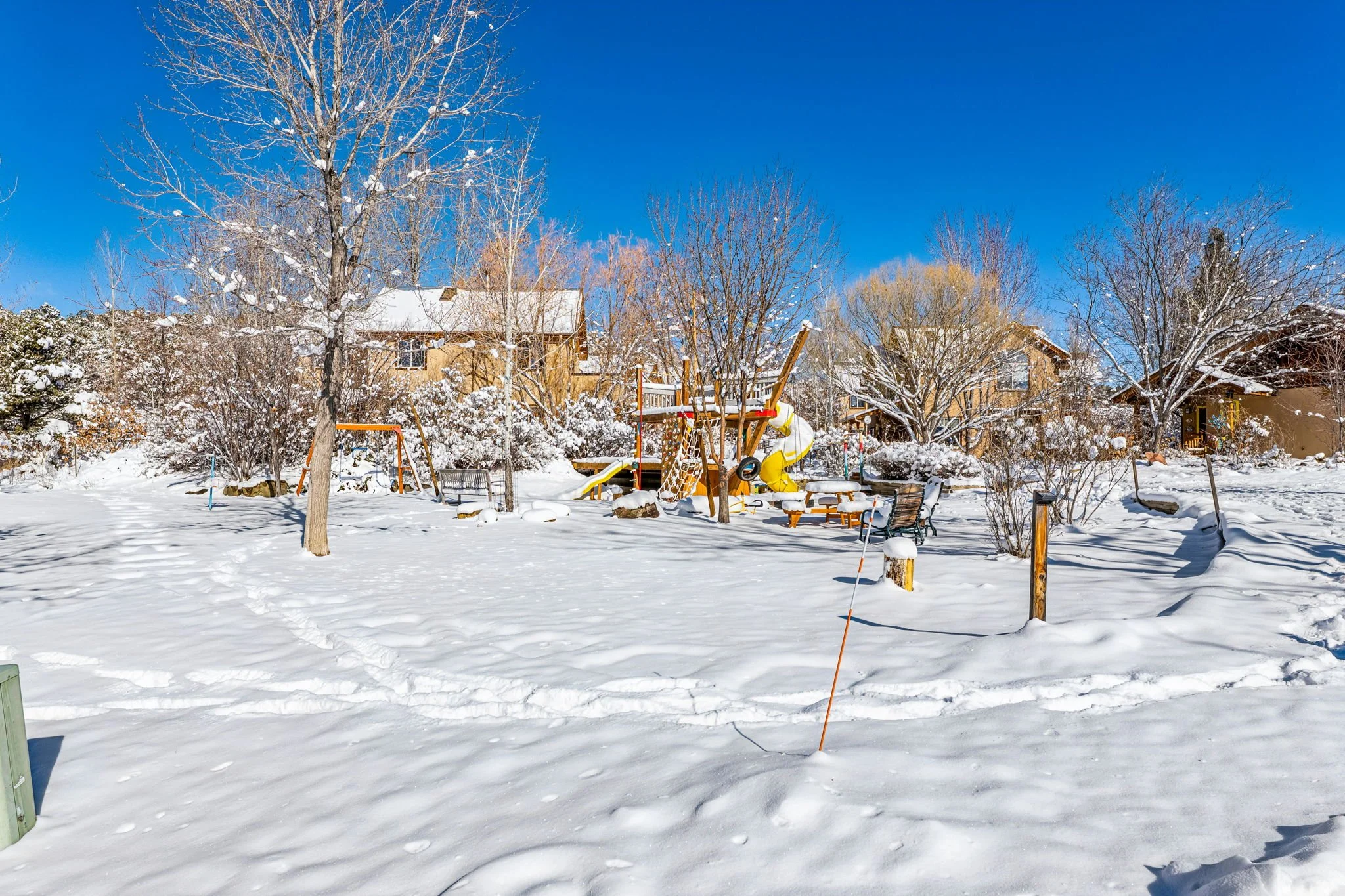Snow-covered backyard with trees, playground equipment, benches, and houses in the background under a clear blue sky.