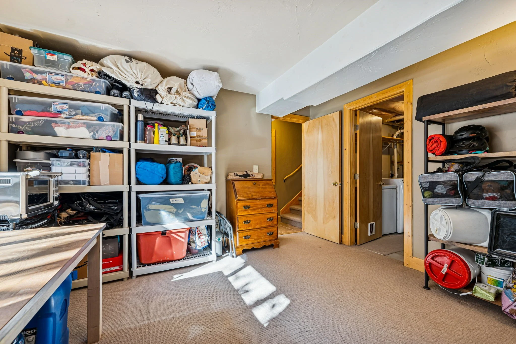 Storage room with shelves filled with boxes, bags, and equipment, small wooden chest, door to a laundry area, and a corner with bins and helmets.