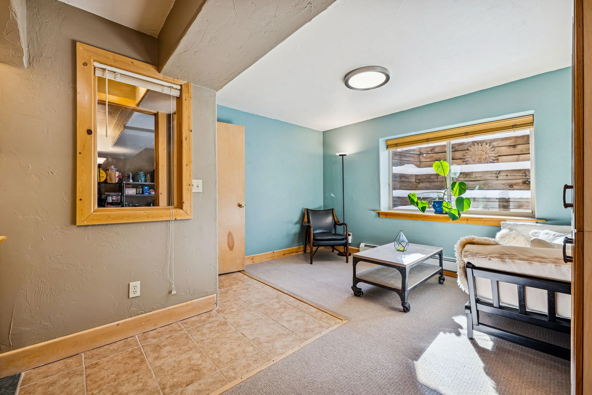 A cozy living room with light blue walls, a window with a wooden frame, a black armchair, a white coffee table on casters, a beige couch with a fur throw, and a tall black floor lamp. There's a houseplant on the windowsill, and a wood-paneled wall ou