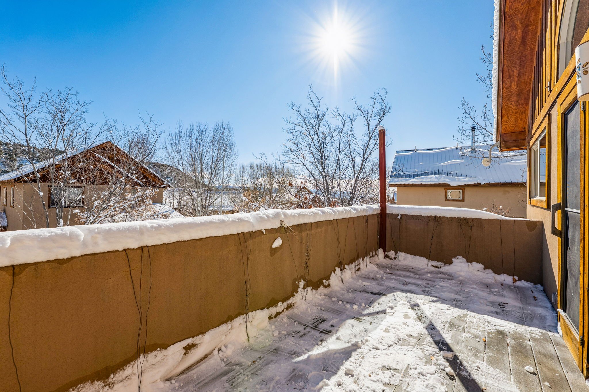 Sunny winter day on a balcony with snow-covered ground and railing, leafless trees and neighboring buildings in the background, clear blue sky, and the sun shining brightly.