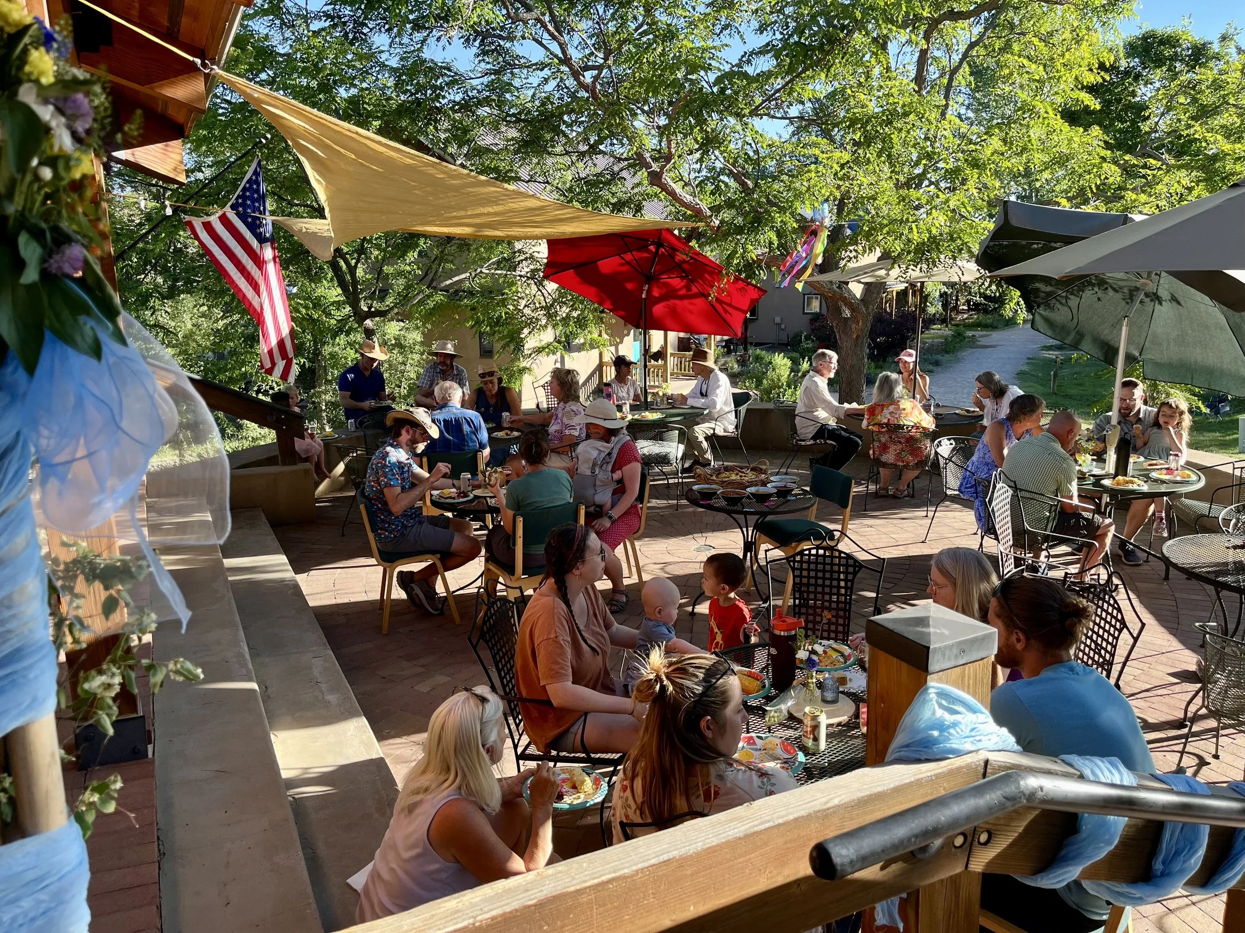 People gathered on a wooden outdoor patio with tables, chairs, colorful umbrellas, and an American flag, enjoying a sunny meal surrounded by trees.