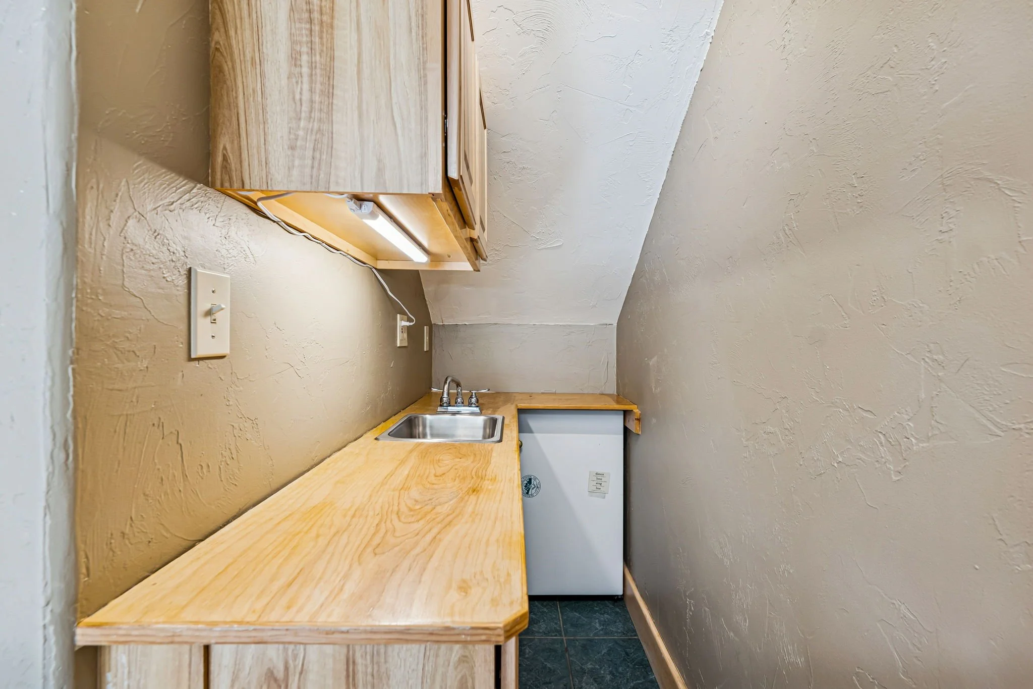 A small kitchenette with a wooden countertop, a stainless steel sink, upper cabinets, and a mini refrigerator beneath the countertop. The walls are beige with a textured finish, and there is a fluorescent light fixture under the cabinets.
