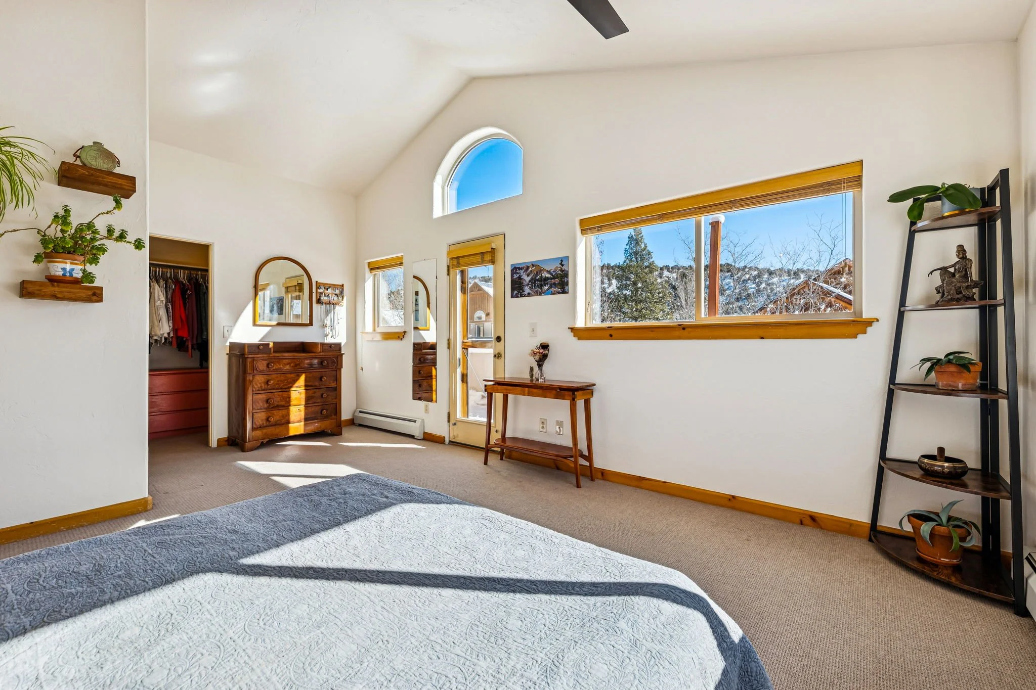 A bright bedroom with white walls, a high ceiling, and large windows showing a snowy outdoor scene. The room has wooden trim, furniture, and a dark metal ladder-style shelf with plants and decorations.