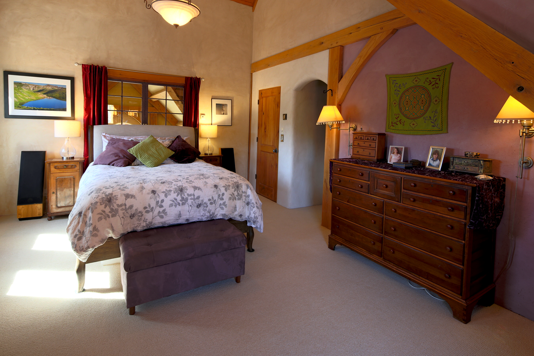 Bedroom with a bed featuring a floral duvet, multiple pillows, nightstands with lamps, a window with red curtains, a wooden dresser with framed photos, a green wall hanging, and beige walls with wooden accents.