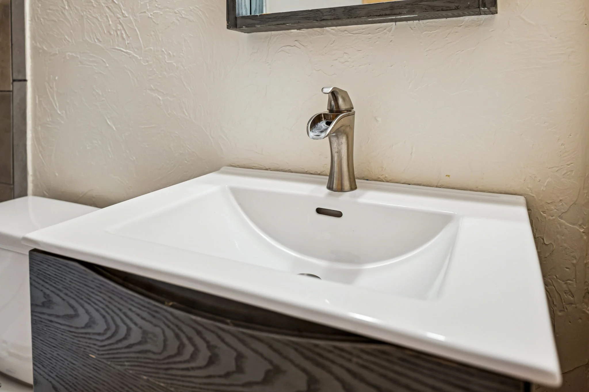 A white bathroom sink with a metallic faucet mounted on a textured beige wall, partially visible mirror above, and part of a black cabinet below.
