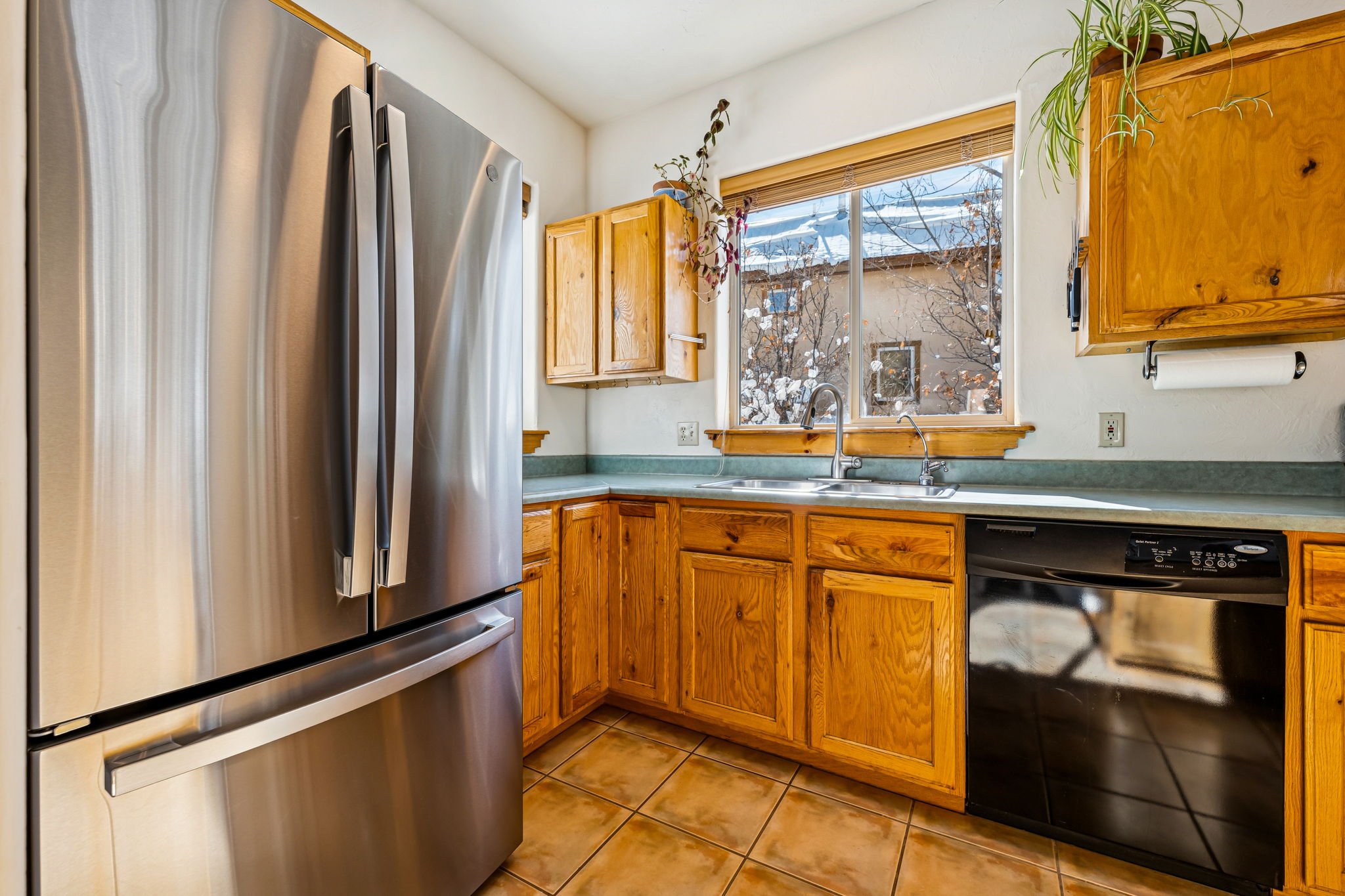 Kitchen with wooden cabinets, a stainless steel refrigerator, a window with a view of snow outside, a black dishwasher, and a green countertop.