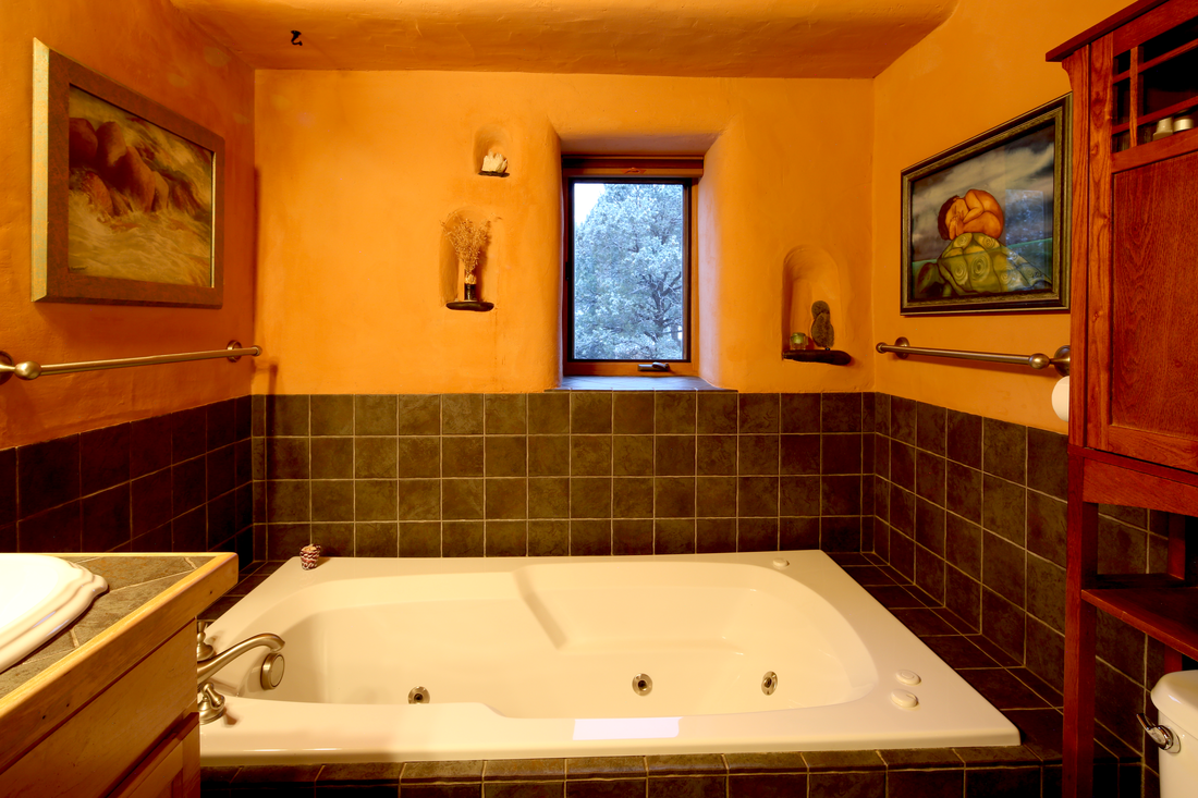 Bathroom with a jetted tub, orange walls, dark green tiles, window showing trees outside, paintings, and wooden fixtures.
